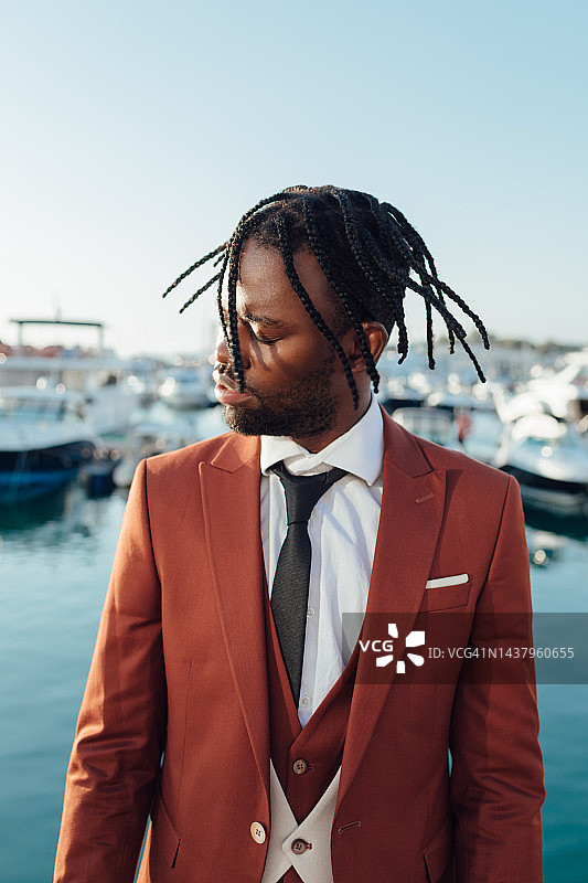 戴着帽子、留着脏辫、穿着棕色西装和白色裤子的黑人青年男子，戴着太阳镜站在大海和游艇港口的背景下，摇着头图片素材