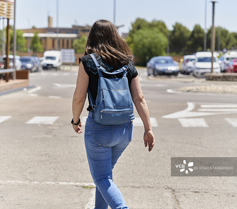 长发女人，穿着牛仔裤，背着背包走在街上，背对着镜头，阳光明媚，有停放的汽车图片素材