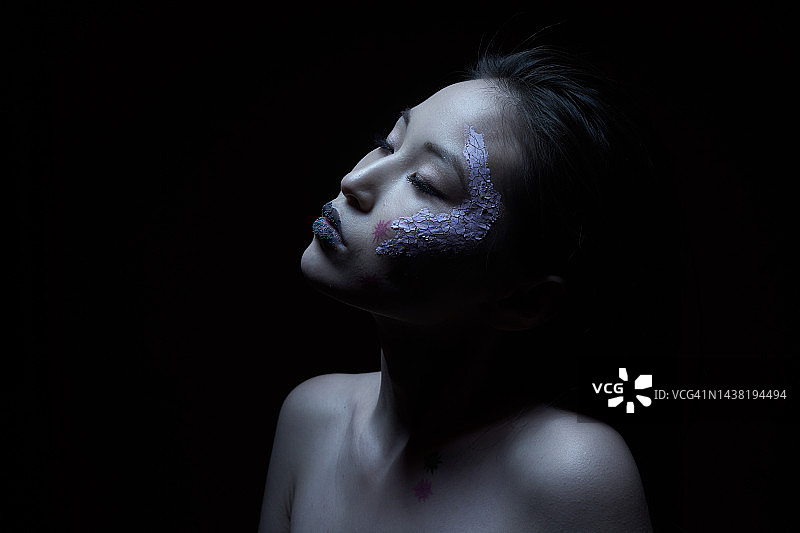 一位年轻的亚洲女性脸颊上贴着粉色面膜，脸上画着病毒，站在黑色背景前看向别处。疾病、感染、心理健康、艺术摄影时尚概念图片素材