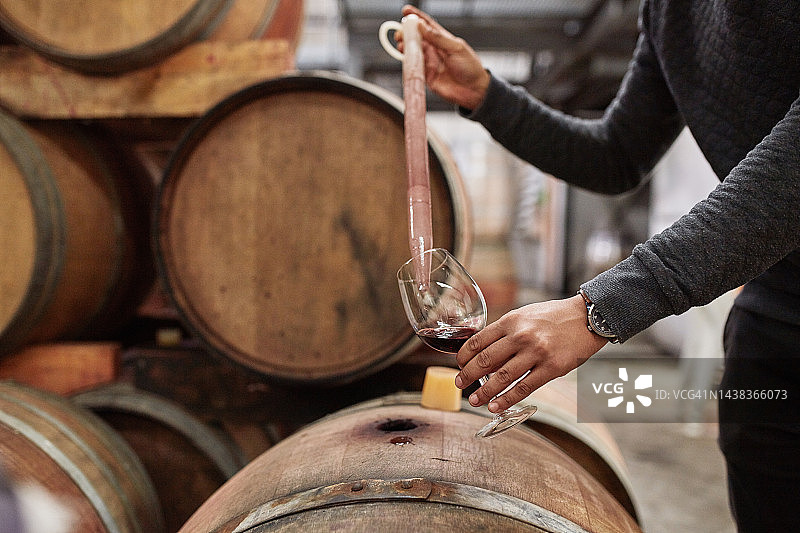 在葡萄酒厂，工人用移液管从木制储酒桶中检测、品尝或取样一杯年份红酒。酒精行业工具，质量控制检查或人员检查液体饮料图片素材