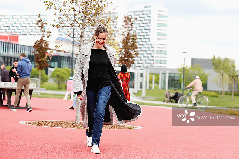 身穿灰色外套的自信年轻女性穿过公共游乐场（正面视角）图片素材