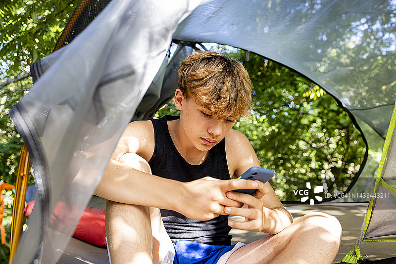 英俊的青少年男孩坐在悬挂的吊床帐篷上使用手机图片素材