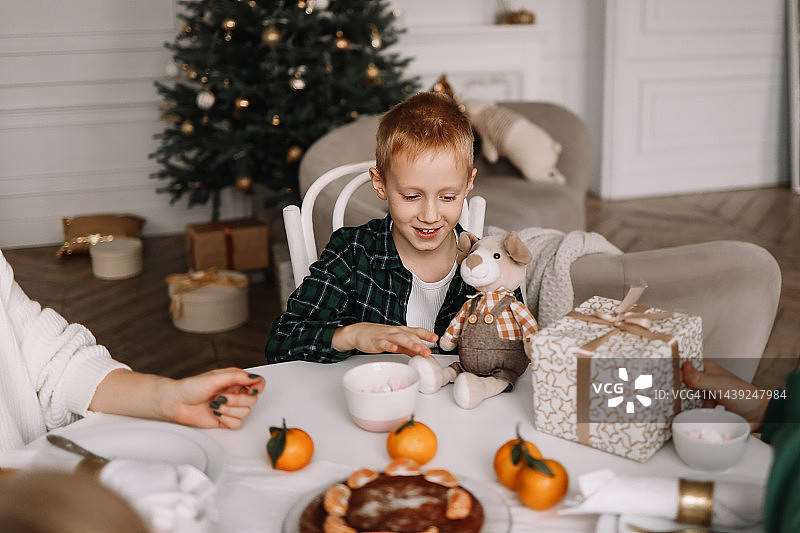 微笑的青少年男孩在装饰过的房间里，圣诞树旁边的餐桌旁拿着毛绒玩具礼物，过圣诞节晚餐图片素材