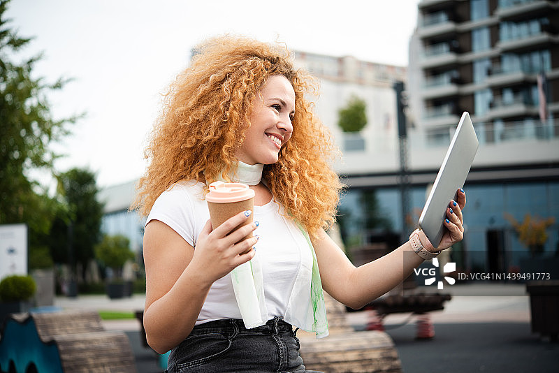 年轻女子在街上手持平板电脑，一边喝咖啡一边进行视频通话图片素材