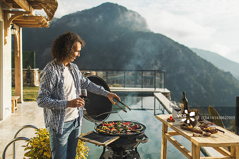 年轻男人在泳池边烹饪烤蔬菜，欣赏山景并品尝葡萄酒图片素材