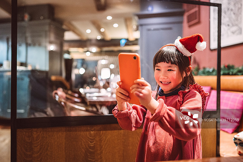 一位戴着圣诞老人帽发带的可爱女孩在餐厅用餐时用智能手机自拍图片素材