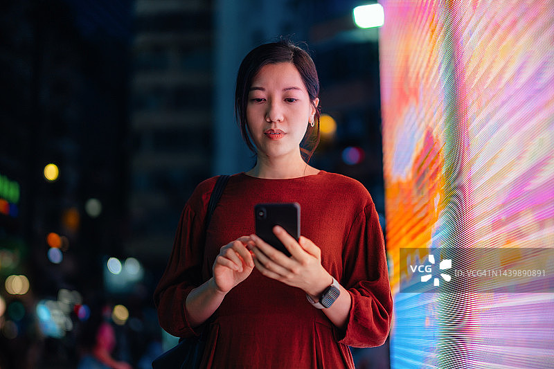 年轻的亚洲女人在夜晚繁忙的城市街道上使用智能手机，站在色彩缤纷的LED数字显示屏前。背景是散景灯光的城市夜景。生活方式与科技：通过科技保持联系图片素材