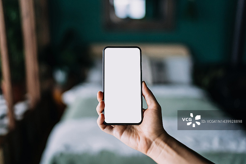特写：舒适时尚卧室里，女性手持空白白色屏幕智能手机。生活方式和科技。带有空白屏幕的智能手机，用于设计模型图片素材