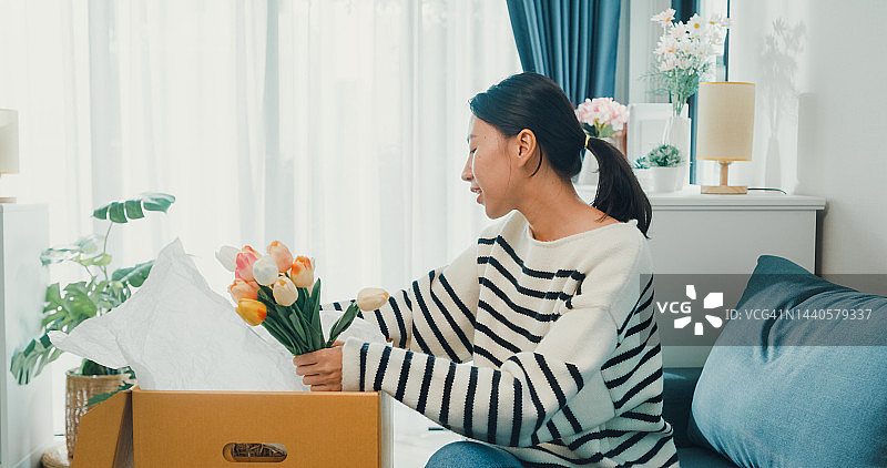 穿着毛衣，从家具店收到并打开纸板箱装饰的兴奋年轻亚洲女孩，在沙发上享受快乐时光图片素材