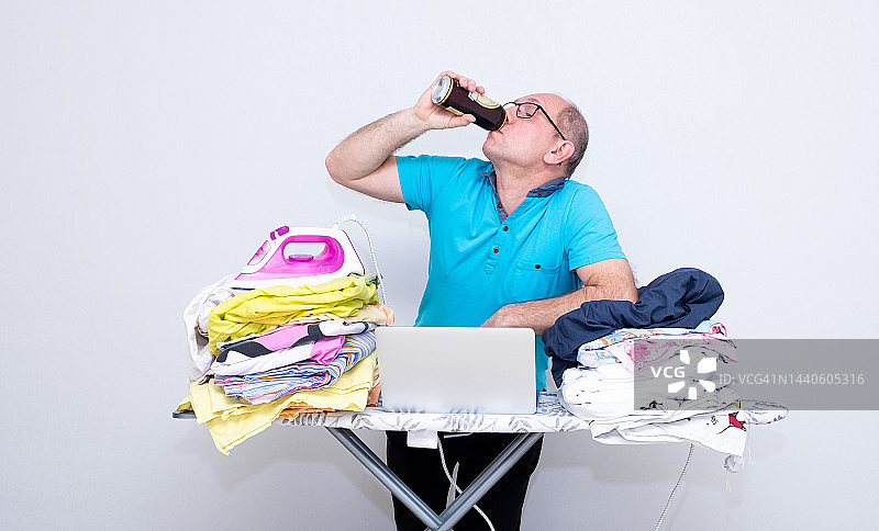 白人男子喝着啤酒，用笔记本电脑计划熨烫床单图片素材