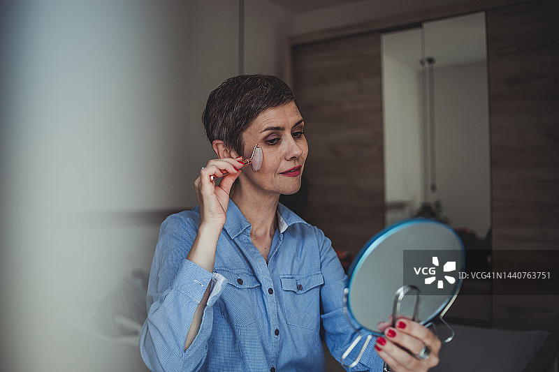 一位留着短发的中年妇女坐在卧室的床上，用 derma roller 按摩她的脸图片素材