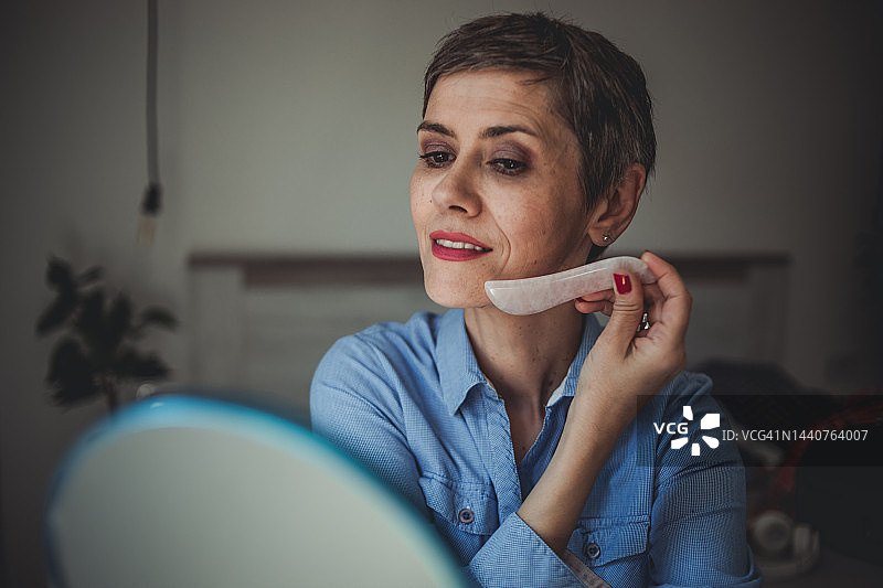 一位留着短发的中年妇女坐在卧室的床上，用 derma roller 按摩她的脸图片素材