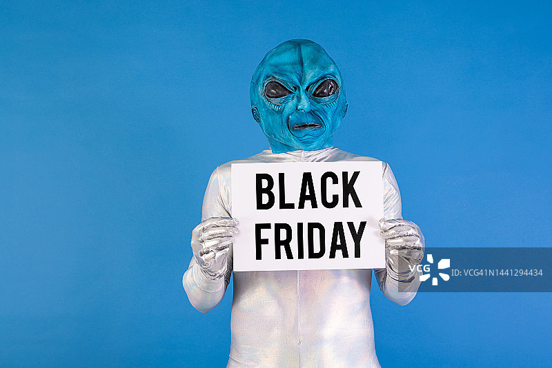 打扮成蓝色外星人的人拿着一个白色标志，上面用黑色写着“黑色星期五”。关于优惠、折扣、机会、有趣、奇怪和外星人的概念图片素材