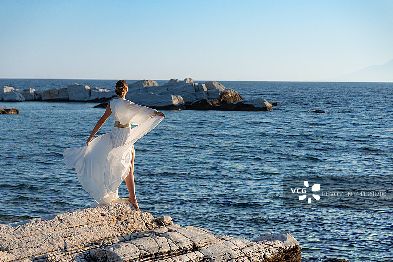 穿着金色腰带的白色无袖长裙的年轻美女在岩石海岸线上玩着丝绸裙摆图片素材