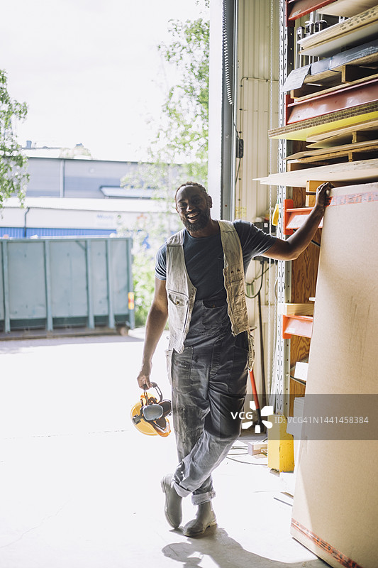 微笑的男性木匠全身照，戴着安全帽，站在仓库的木板旁。图片素材