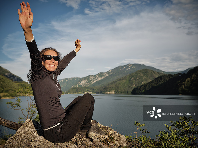 快乐的女人在湖边的岩石上举起双臂图片素材
