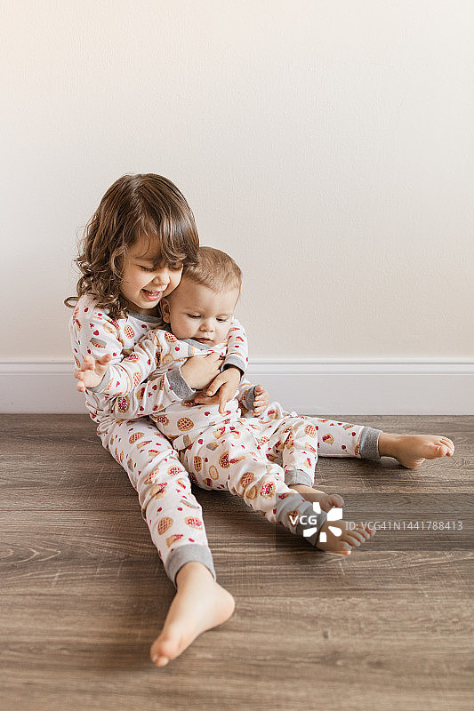 三岁幼儿女孩坐在地板上抱着她10个月大的弟弟，他们穿着相配的樱桃派睡衣，在2022年11月在家中度过温馨的感恩节图片素材