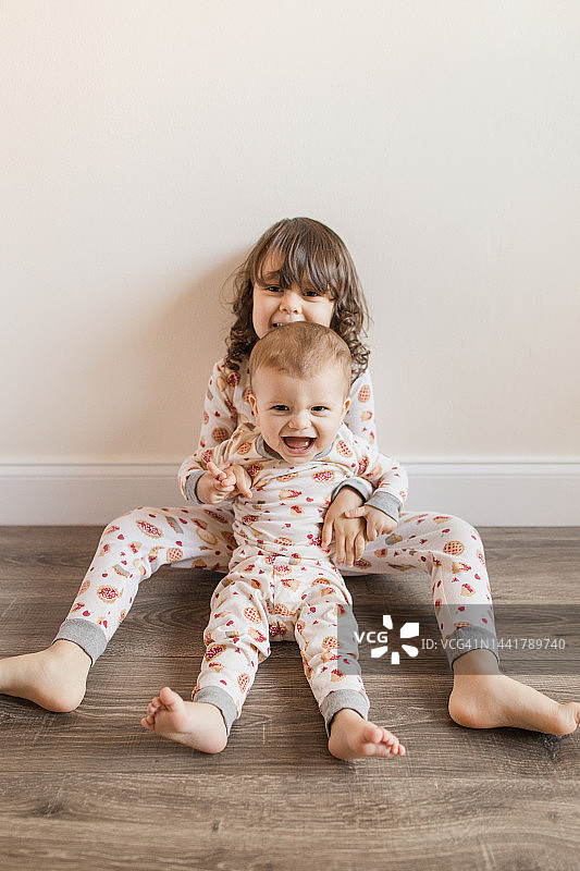三岁幼儿女孩坐在地板上抱着她10个月大的弟弟，他们穿着相配的樱桃派睡衣，在2022年11月在家中度过温馨的感恩节图片素材
