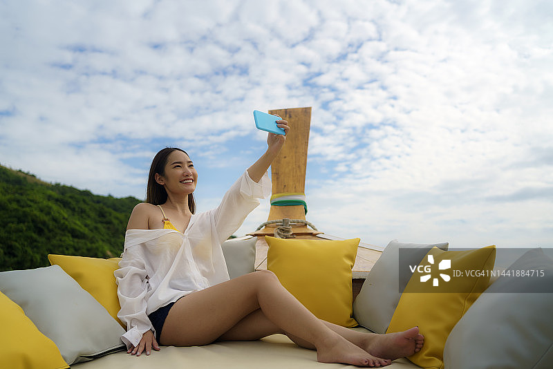 在泰国普吉岛夏季假期期间，亚洲女人坐在长尾船上用手机自拍图片素材