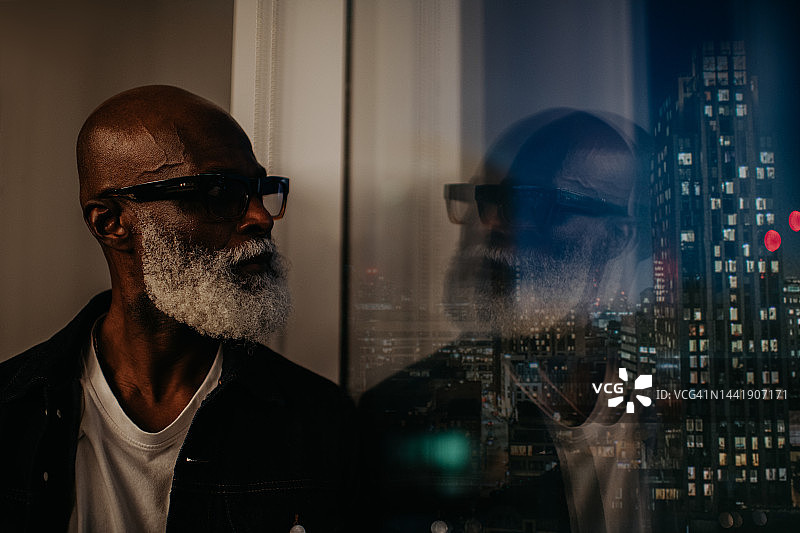一位沉思的老年男人站在窗前，望着夜晚的城市天际线特写图片素材