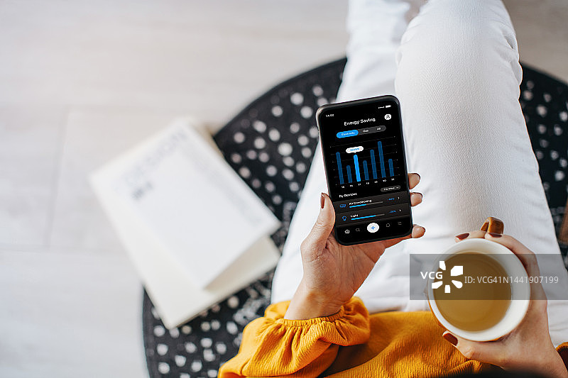 年轻的亚洲女性在家中使用智能手机上的智能家居能源监控应用程序，查看和管理家庭能源使用情况。能源控制和效率管理概念图片素材