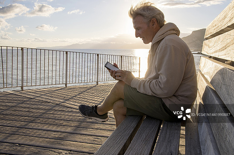 日出时，在海边长椅上放松地看电子书的中年男人图片素材