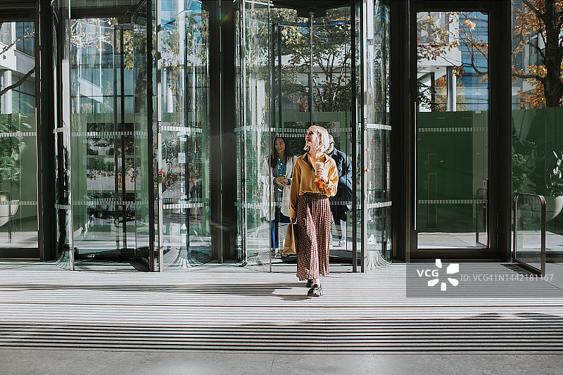 一位女士手拿咖啡和外卖纸袋，通过旋转门进入商业大楼图片素材