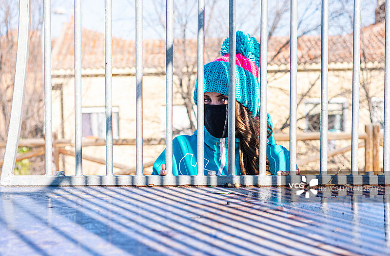 戴着面罩、身穿蓝色外套和蓝色羊毛帽的女孩在铁栅栏后的肖像图片素材