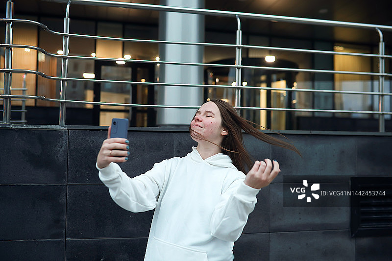 女子在玻璃金属办公楼前自拍，身穿白色运动帽衫图片素材