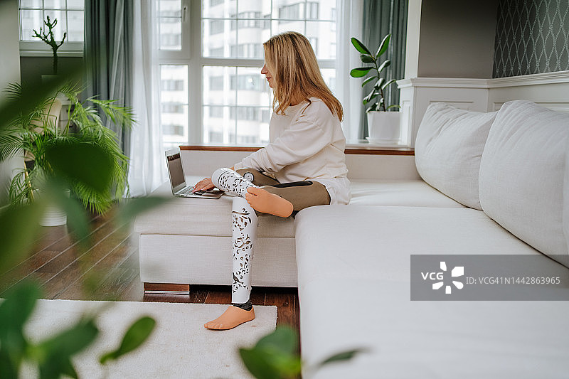 使用笔记本电脑在客厅工作的安装假肢的金发女人图片素材