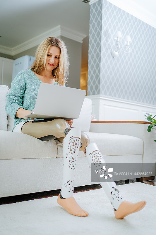 在家办公的IT支持。使用笔记本电脑的金发女性（装有人工腿），在客厅工作图片素材