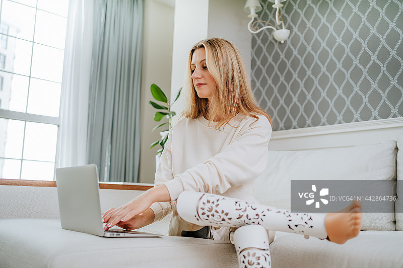 使用笔记本电脑在客厅工作的安装假肢的金发女人图片素材