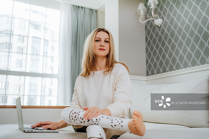 在家工作的女程序员：使用笔记本电脑的金发假肢女性在客厅工作并看向镜头图片素材