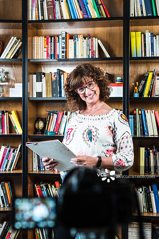 中年教师用摄像机录制在线课程，背景是图书馆和书籍图片素材