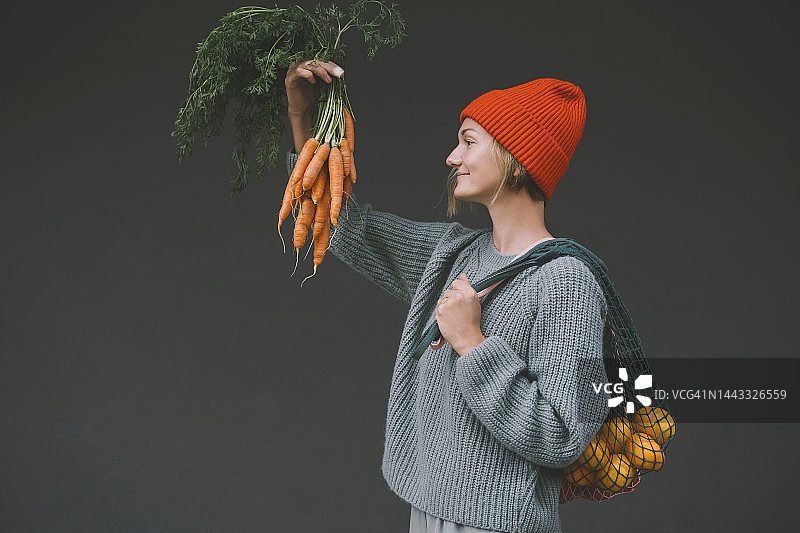 携带网状袋，在灰色墙前看胡萝卜的女人图片素材