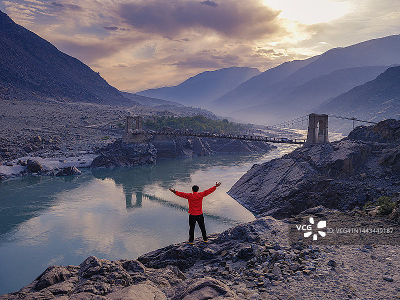 旅行者身穿红色外套站在河边的岩石悬崖上，背景是拉科特桥的交通道路和桥梁，位于巴基斯坦北部吉尔吉特-巴尔蒂斯坦的喀喇昆仑公路图片素材