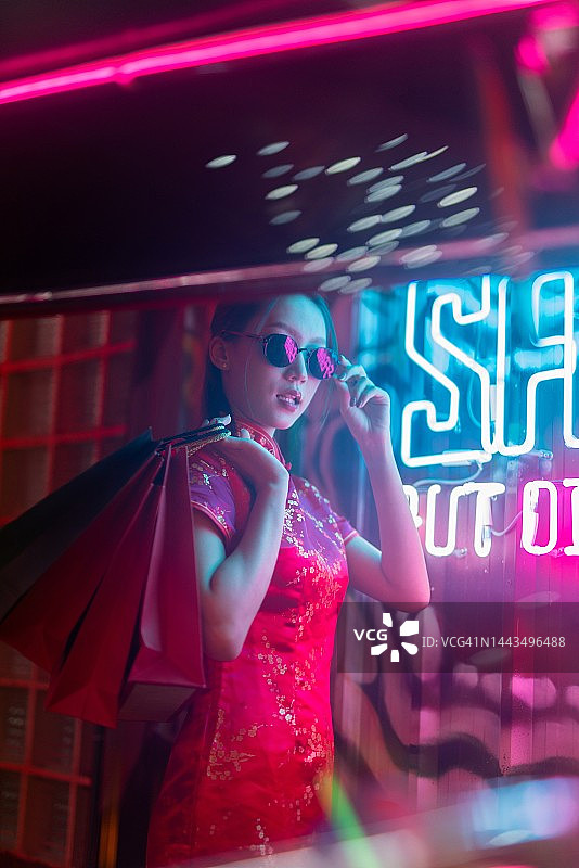 穿着红色中式旗袍、戴着太阳镜的年轻亚洲女性在霓虹灯前手提购物袋图片素材