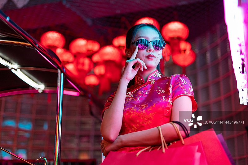 年轻亚裔女模特身穿红色中式旗袍，在霓虹灯前摆酷，手提购物袋图片素材