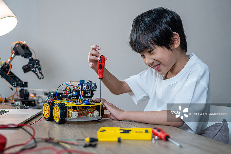 戴眼镜的小书呆子拿着一个机器人。一个穿着灰色衬衫戴眼镜的男孩坐在桌子前，桌子上是他制作的机器人。他对自己的工作非常满意图片素材