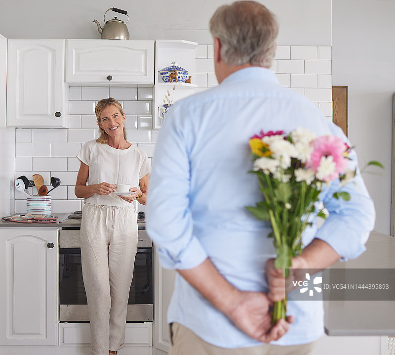 老年夫妇在厨房里收到鲜花惊喜，爱和浪漫。女人、男人和退休一起拿着花束庆祝婚姻、生日或周年纪念日图片素材