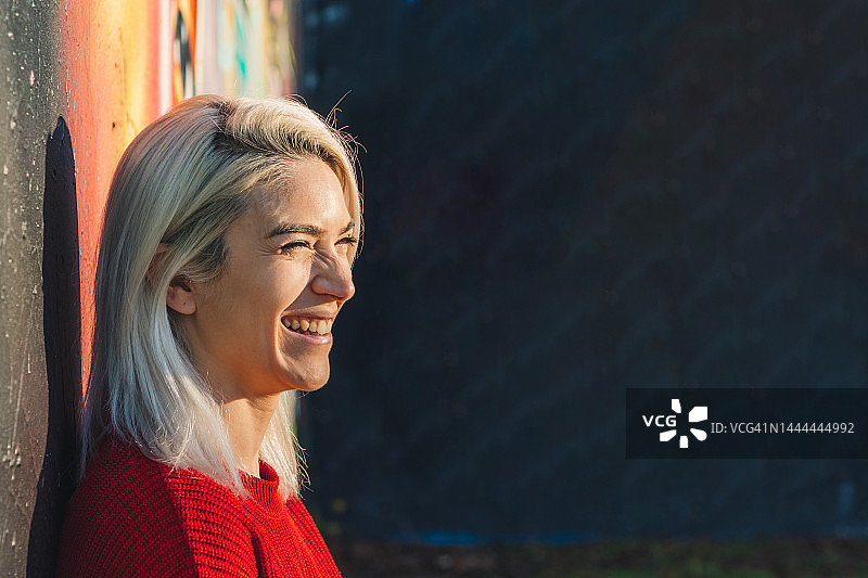 银发女孩靠在涂鸦墙上微笑，身穿红色毛衣图片素材