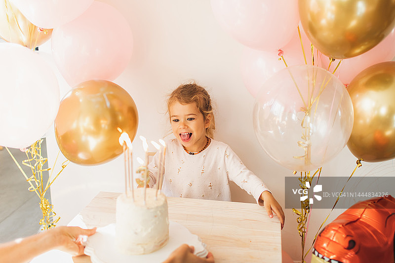 可爱的小女孩在家中吹灭生日蛋糕上的蜡烛，背景是气球。图片素材