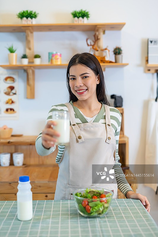 年轻亚洲女人穿围裙在厨房喝牛奶吃沙拉图片素材