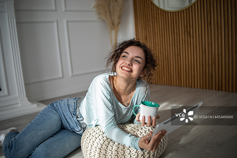 年轻迷人的微笑女性穿着休闲家居服坐在地板上喝早咖啡，使用平板电脑图片素材
