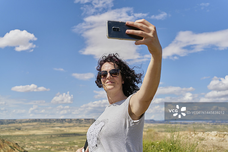 女人戴着墨镜在户外用手机自拍，背景是蓝天白云图片素材