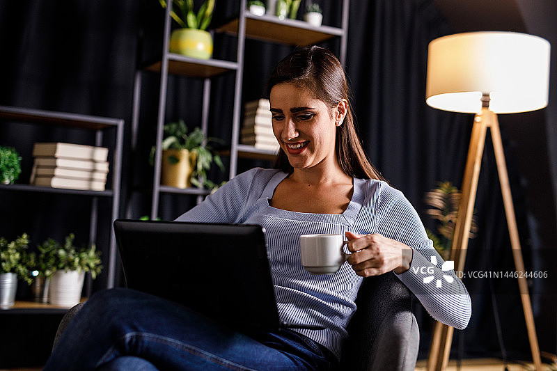 快乐的年轻女子在扶手椅上放松，下班回家后使用笔记本电脑图片素材