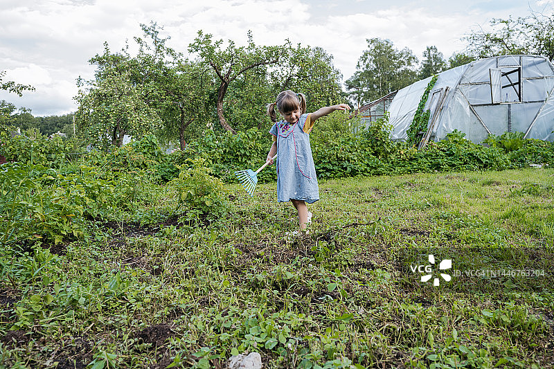 学龄前女孩穿着蓝色连衣裙站在花园中央，在阴天里手里拿着一把小耙子图片素材