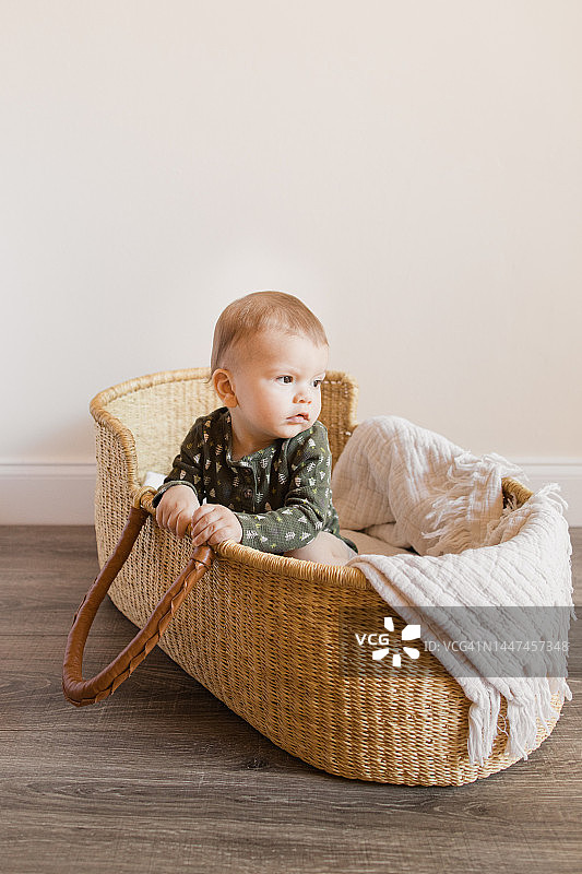 2022年12月，一位11个月大的圣诞男婴穿着带有小圣诞树的深绿色保暖连体衣，在地板上的海草摩西篮里图片素材