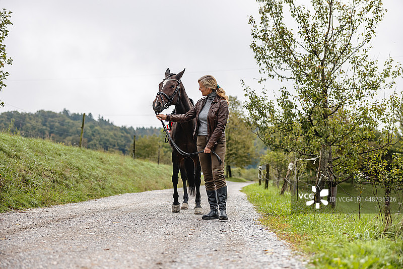 快乐的成年白人金发女性在乡村公路的马旁撑伞图片素材