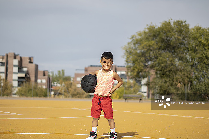 可爱的拉美裔男孩站在室外公园的篮球场上，拿着篮球，肖像 - 健康的生活方式 - 返回学校图片素材